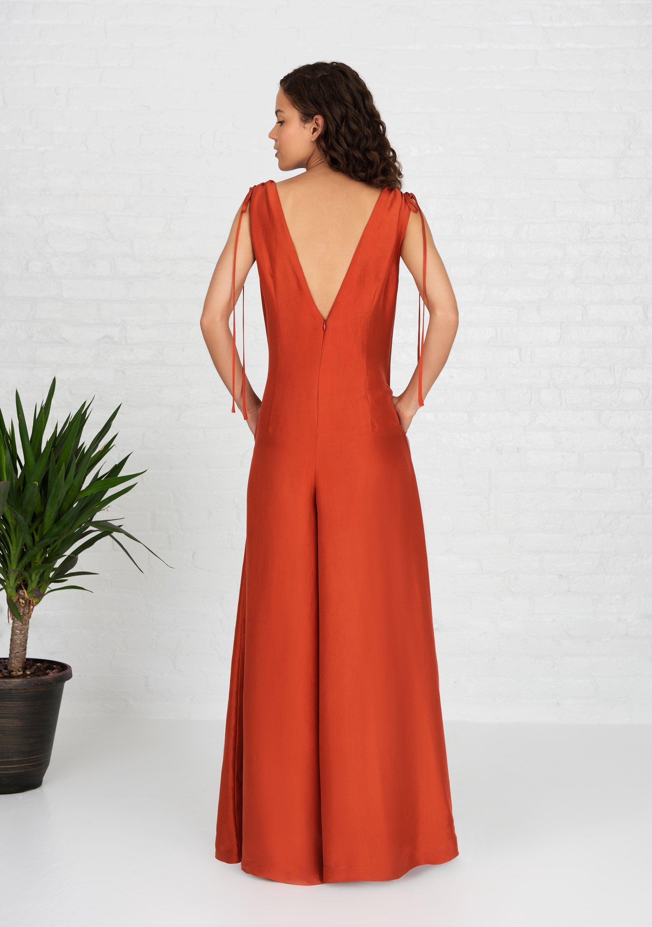 back profile view of woman wearing burnt orange sleeveless silk jumpsuit with hands in her pockets