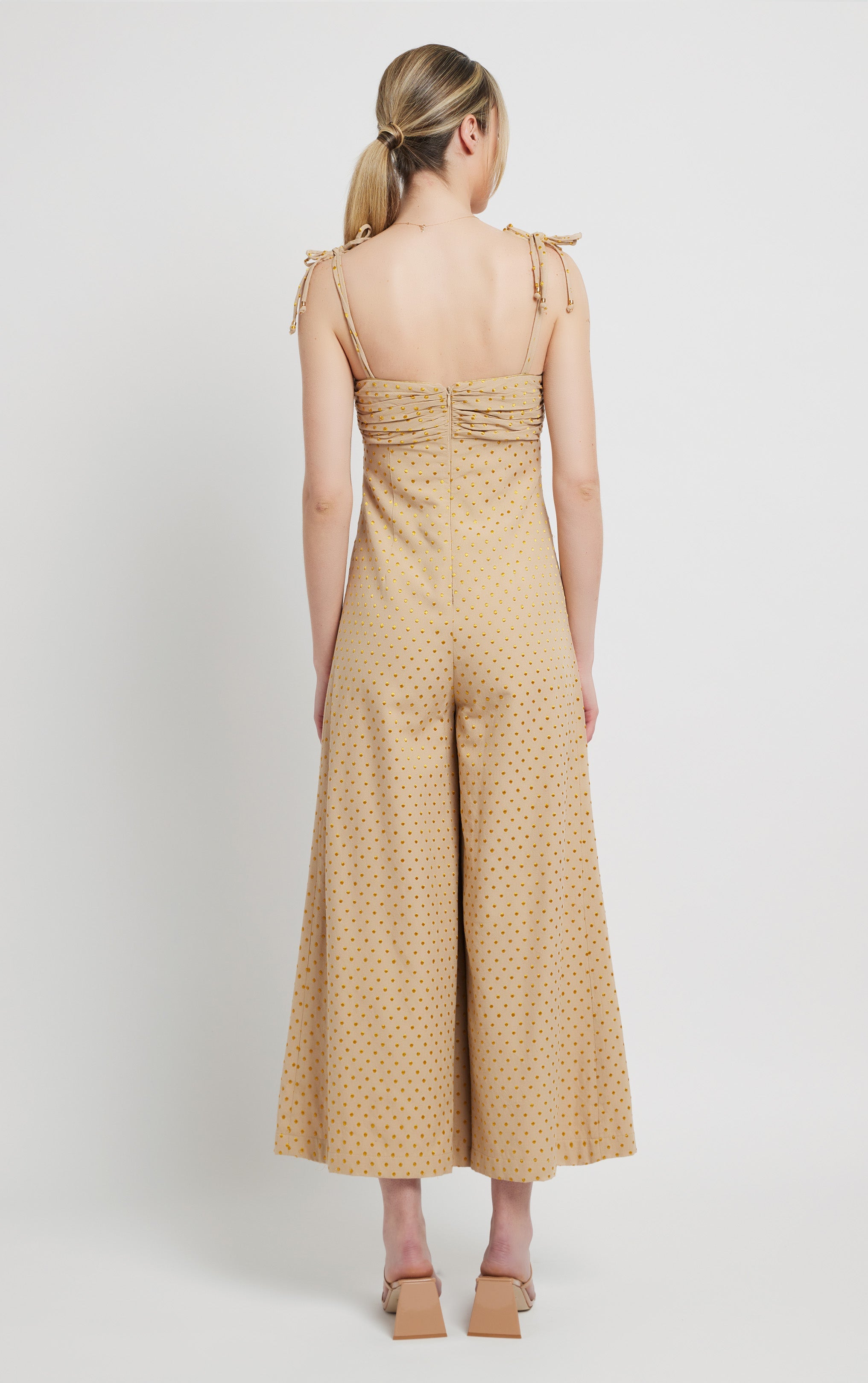 back profile of woman standing wearing khaki sleeveless jumpsuit with gold polka dots