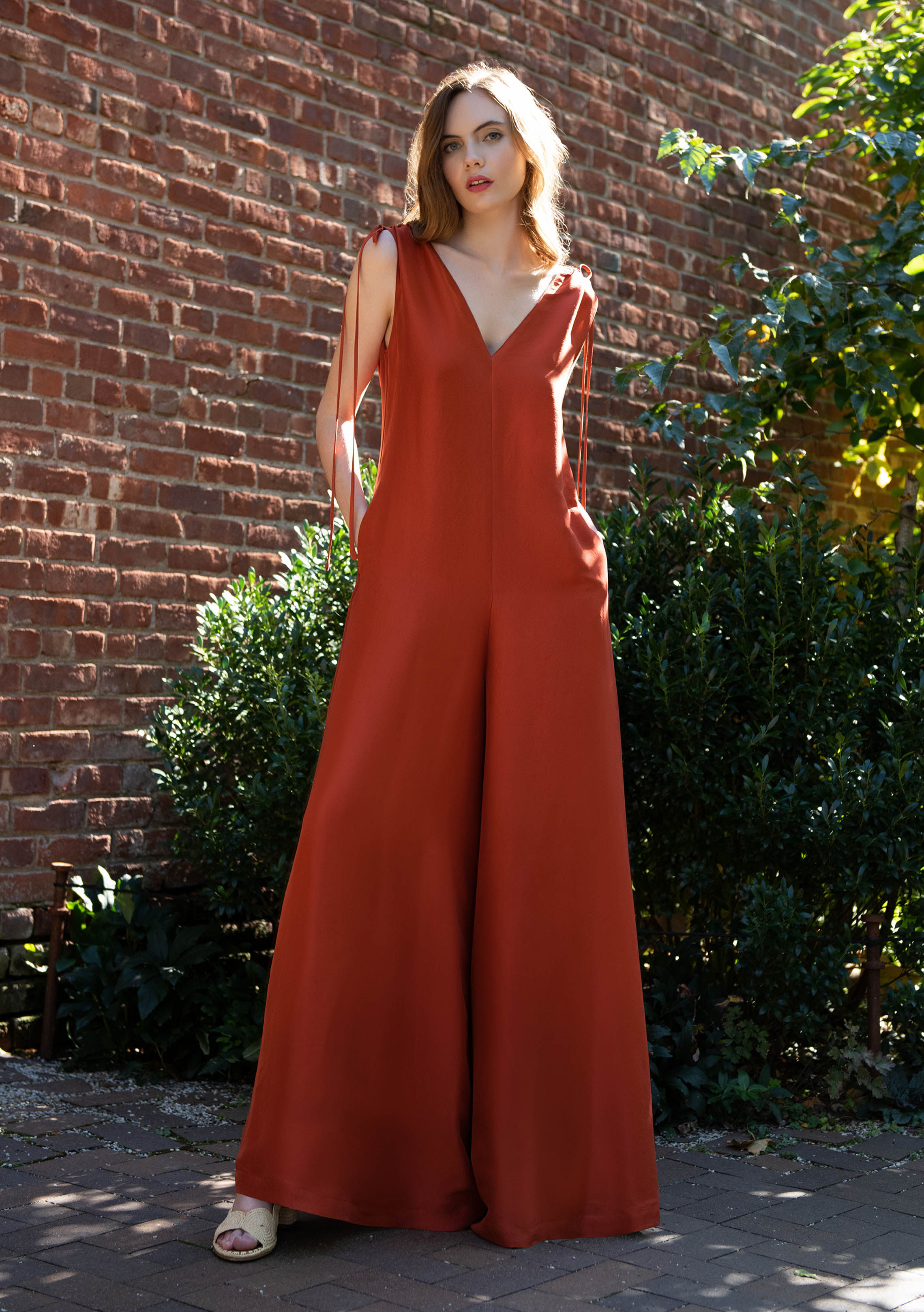 woman wearing burnt orange sleeveless silk jumpsuit with hands in her pockets